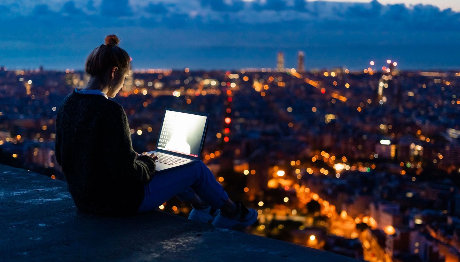 Persona trabajando en un portátil frente a una ciudad iluminada al anochecer